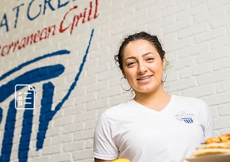 A woman in a white shirt stands smiling in front of a brick wall with the words "Mediterranean Grill" painted on it. She is holding a plate of food, representing a popular Greek fast food franchise.