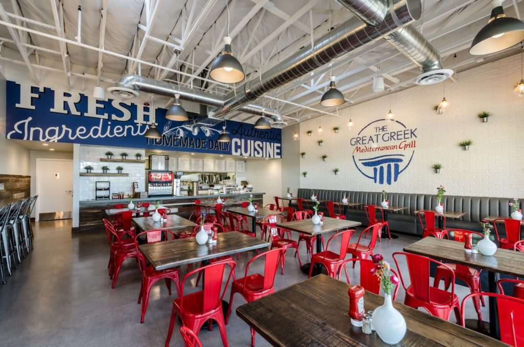 Interior of a restaurant with a modern design, featuring red chairs and wooden tables. The walls display the words "Fresh Ingredients" and "The Great Greek Mediterranean Grill," reflecting the quality of this Greek food franchise.