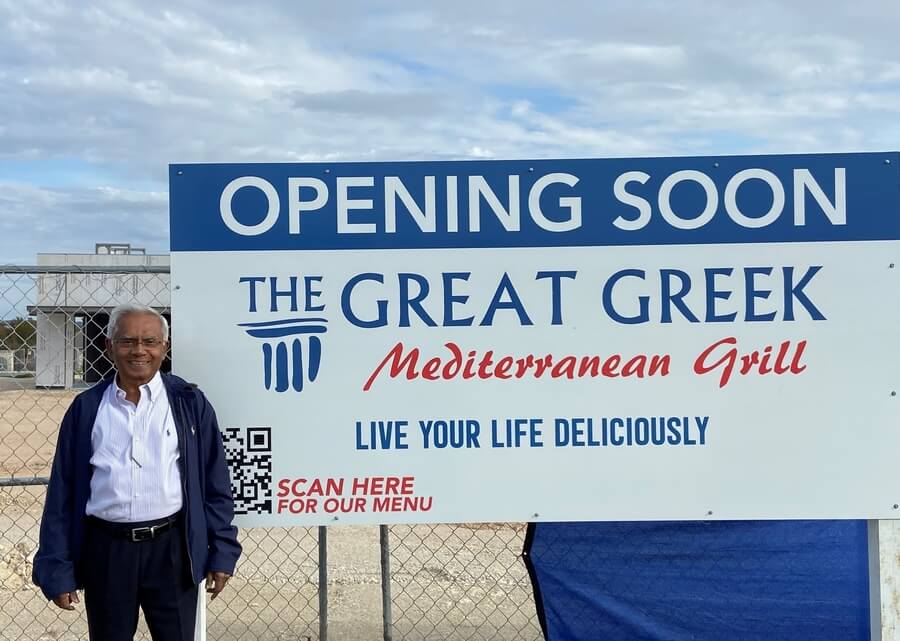 A man stands next to a large sign that reads "Opening Soon - The Great Greek Mediterranean Grill - Live Your Life Deliciously." A QR code is visible on the sign. This new Greek fast food franchise promises to deliver a delightful culinary experience.
