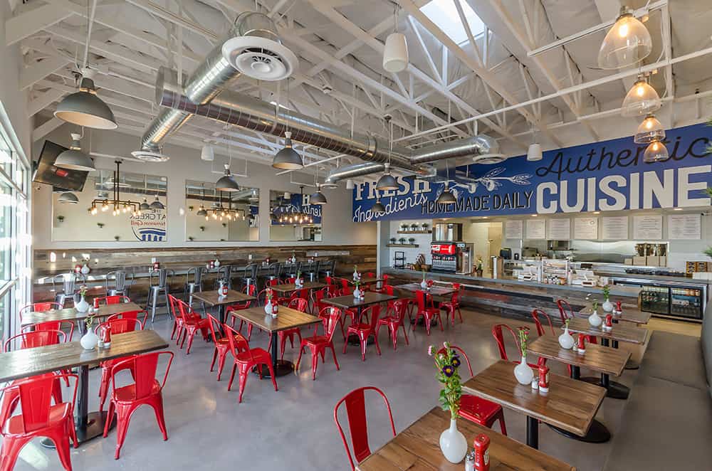 A modern, spacious restaurant with red chairs, wooden tables, and an exposed ceiling. The wall displays "Fresh Ingredients" and "Authentic Cuisine." The counter and kitchen are visible. Perfect for enjoying a quick bite at this Greek fast food franchise.
