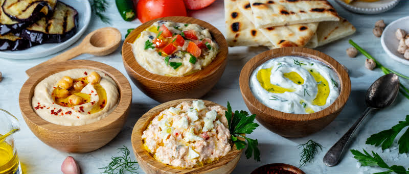 A variety of dips in wooden bowls, including hummus, tzatziki, and other spreads from a popular Greek fast food franchise, are arranged on a table with flatbreads, grilled vegetables, tomatoes, and herbs.