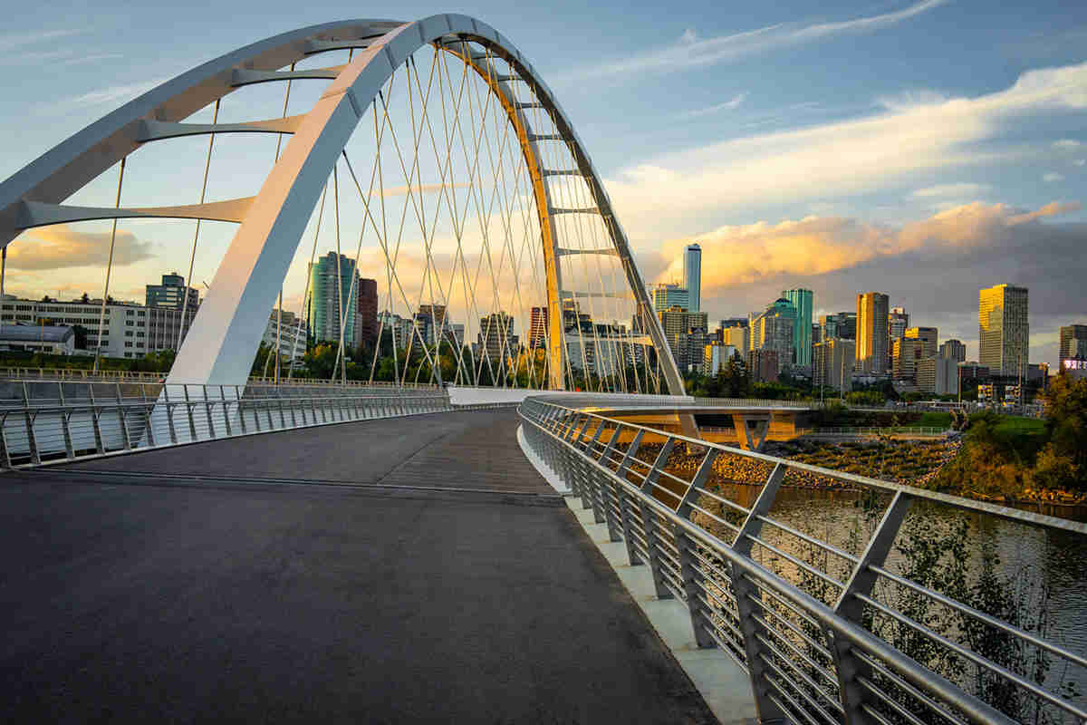 A modern white arch bridge curves over a river, leading towards a city skyline at sunset with tall buildings and a partly cloudy sky. A modern white arch bridge curves over a river, leading towards a city skyline at sunset with tall buildings and a partly cloudy sky.