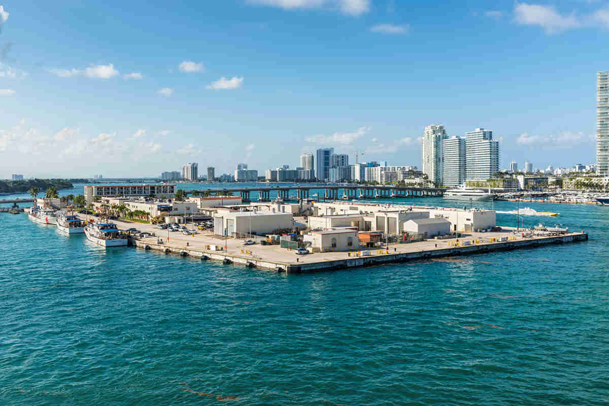 A waterfront area with several buildings and boats, surrounded by water, with city skyscrapers in the background under a clear blue sky.