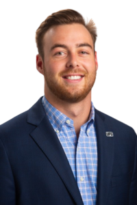A man in a blue blazer with a UFG pin and a checkered shirt, smiling at the camera against a white background.